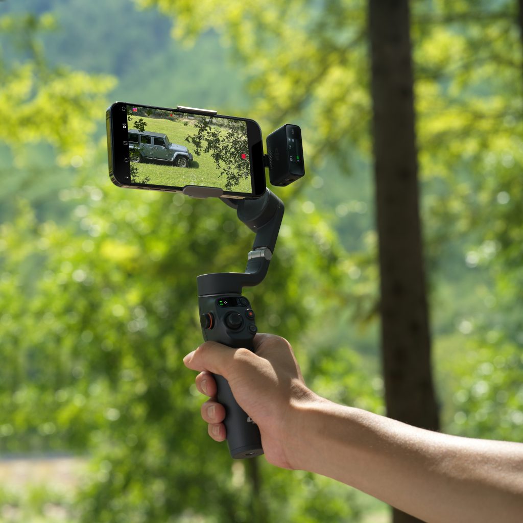 Männerhand hält Smartphone-Gimbal von DJI vor grünem Wald-Hintergrund