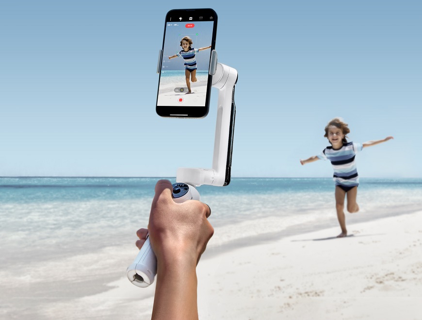 Hand hält Smartphone-Gimbal vor laufendem Kind am Strand
